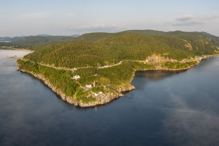 Une vue aérienne d’une eau bleue calme entourant une péninsule vallonnée arborée qui s’étend au loin. 