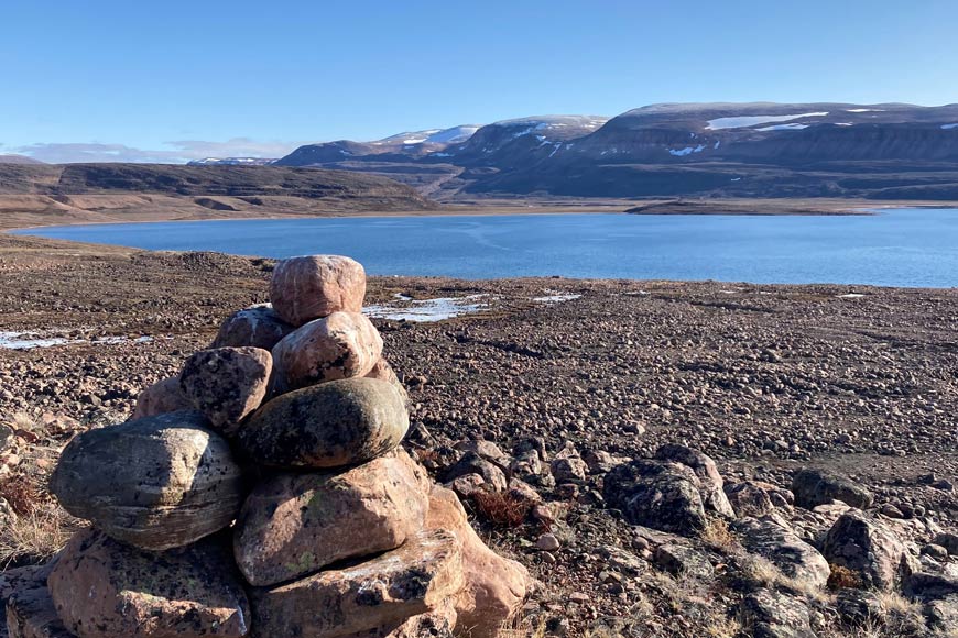 An inuksuk/cairn in the foreground of an Arctic inlet, with low lying snow-capped mountains in the distance.