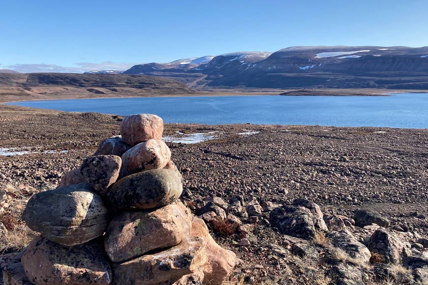 Un inuksuk/cairn au premier plan d’une crique dans l’Arctique, et des montagnes enneigées de faible altitude au loin.