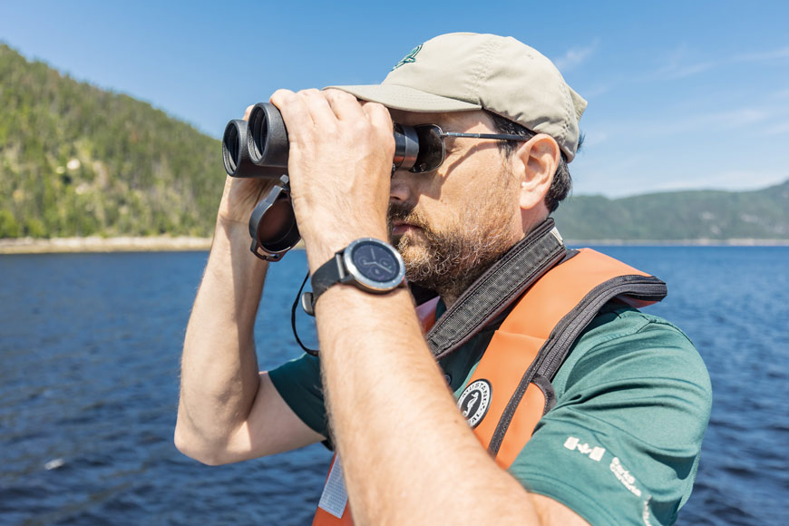 A staff member in Parks Canada uniform on a boat looks through their binoculars out over the water.