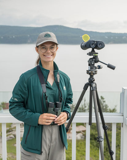 A Parks Canada staff member holds binoculars in front of a fence overlooking a waterbody and a large hill.
