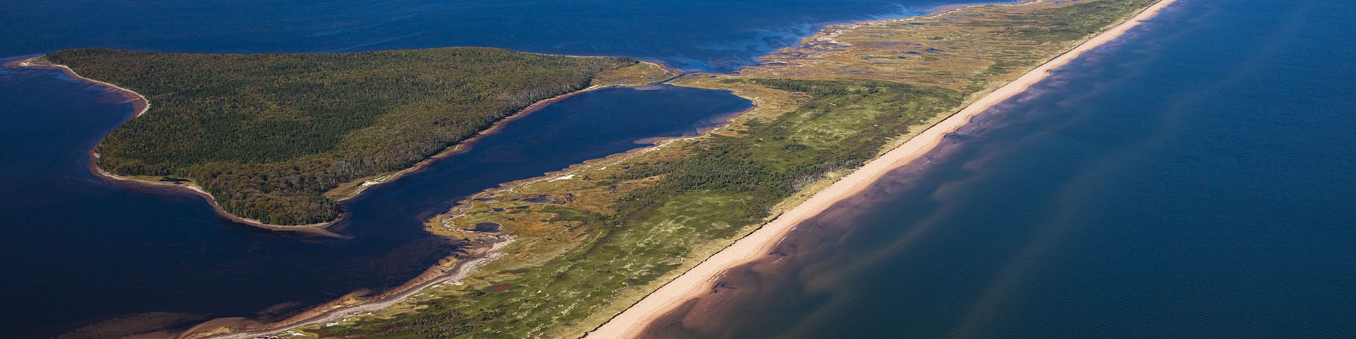 Photo aérienne de la vue de l’ouest sur toute la longueur des dunes Sandhills. L’eau est de couleur bleu nuit et les vagues balayent le sable. La marée monte sur le côté gauche des dunes.