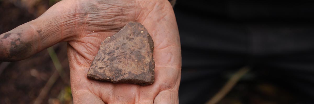A brown ceramic fragment is held in the palm of a hand. 