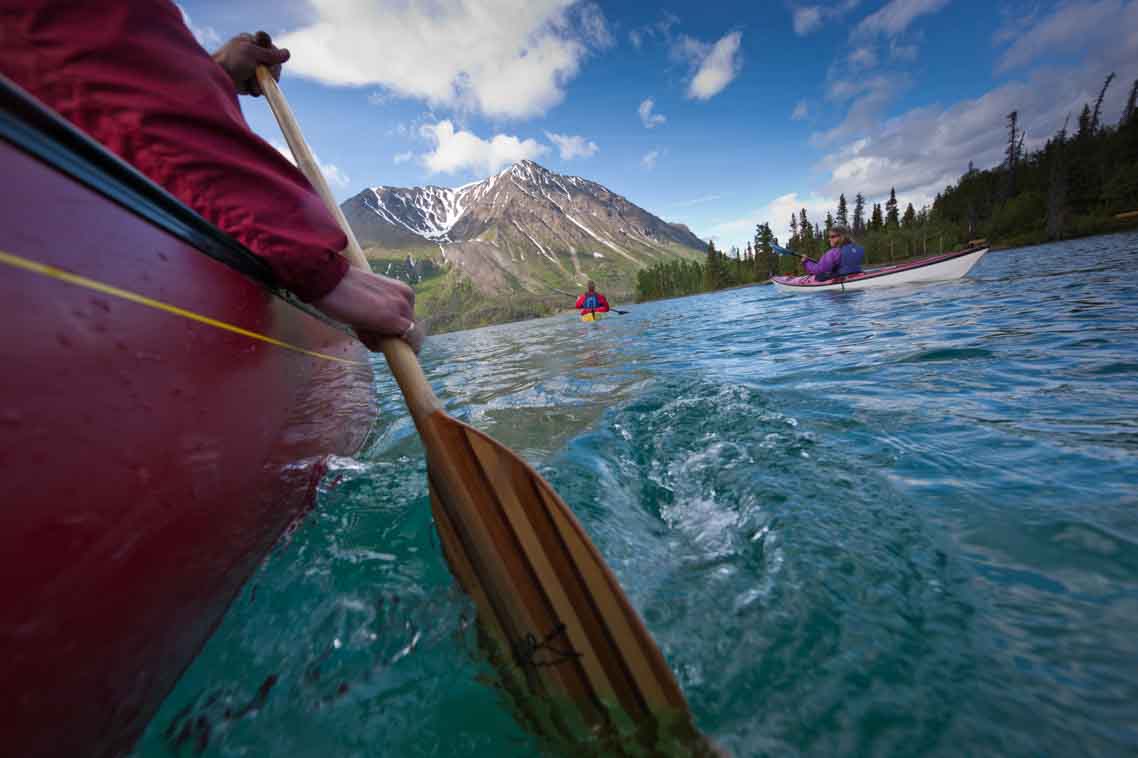 Une personne pagayant dans un canoë rouge avec deux kayakistes devant, une forêt sur la rive à droite et une montagne au loin devant. 