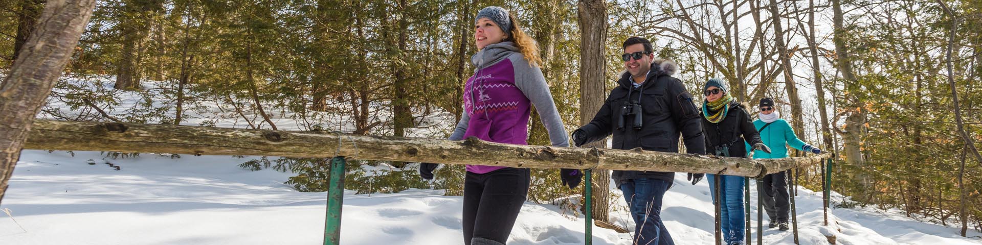 Quatre personnes marchent en forêt dans la neige, ils marchent sur un sentier en se tenant sur un garde.