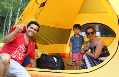 Un adulte fait le signe de la paix tout en souriant, tandis qu'un autre adulte et un enfant sourient, assis dans une tente jaune.
