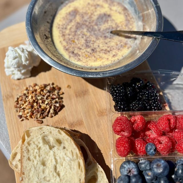 Ingredients displayed on a wooden cutting board, including bread slices, nuts, yogurt, berries, and a bowl of the egg mixture.