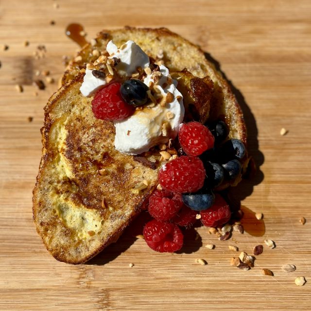 Slices of golden French toast on a wooden board topped with yogurt, fresh fruit, and a drizzle of maple syrup.