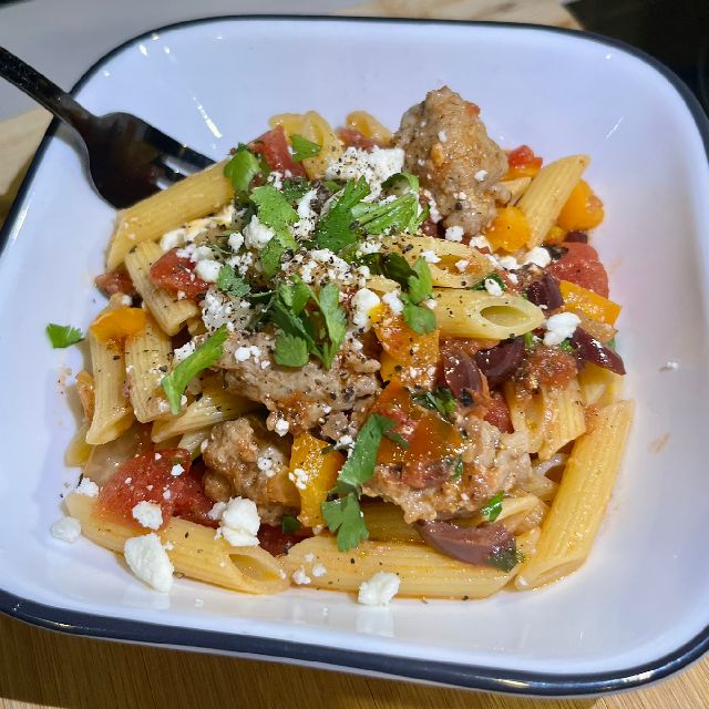  A bowl of penne pasta topped with sausage pieces, Kalamata olives, feta cheese, tomato sauce, and peppers.