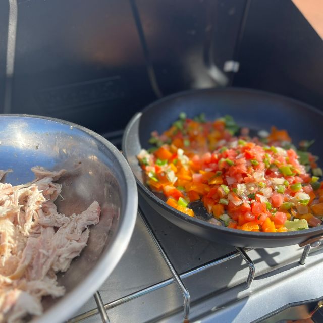 A pan of chopped peppers and onions sit on a camping stove, with a bowl of precooked shredded chicken beside it.
