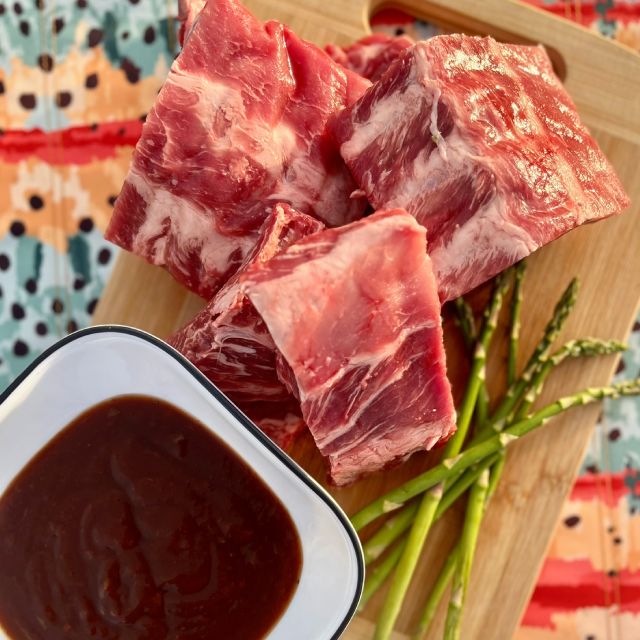 Raw ribs on a wooden cutting board with asparagus spears and a bowl of homemade barbecue sauce beside them.