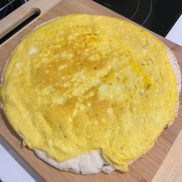 A tortilla laid flat on a wooden cutting board topped with a thin layer of cooked eggs and melted cheese.