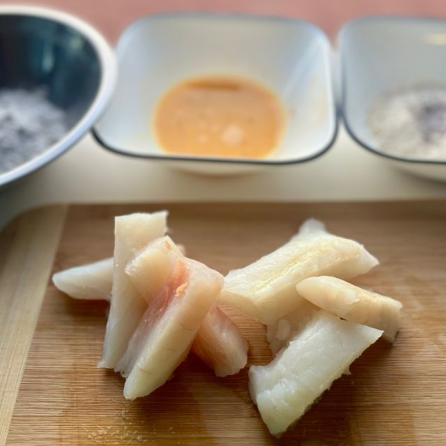 Raw white fish on a cutting board with three bowls behind it containing seasoned flour, beaten eggs, and breadcrumbs.