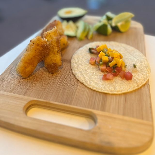 Tacos filled with toppings on a cutting board. Fried fish sits beside them with lime wedges and avocado slices.