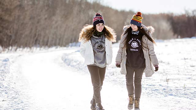 Two woman walk in a snowy forest and smile at each other while wearing their official merchandise.