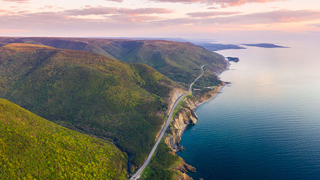 A pink sunset over grassy hills and the cabot trail road by a vibrant blue sea in  Cape Breton Highlands.