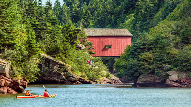 Two people kayak on bright blue waters in front of a red bridge building nestled between trees in Fundy.