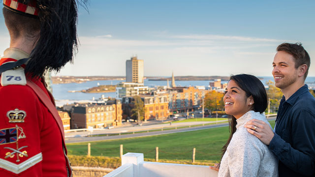Two people speak to a Parks Canada interpreter dressed in a historic outfit, with a historic fort and the sea in the background at the Halifax Citadel.
