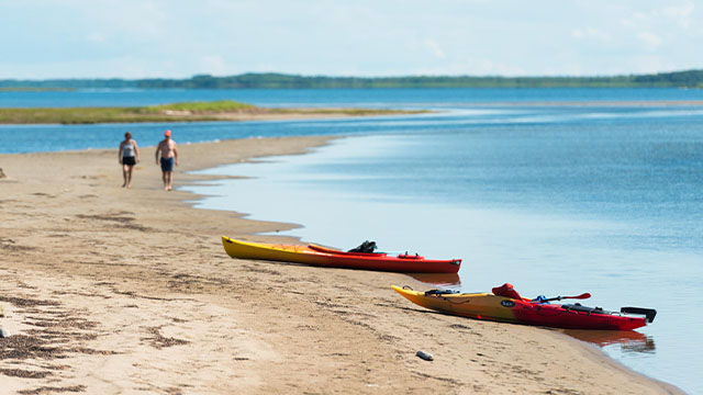 Two kayaks are left on the shore in the foreground, and two people walk on a white sandy beach with bright blue waters in the background at Kouchibouguac.