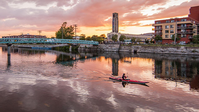 A person kayaks under a pink sunset with clouds and buildings reflecting on Lachine Canal waters.