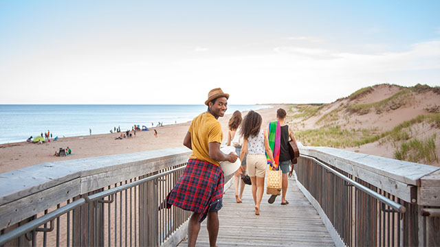 A man turns and smiles at the camera while walking on a beachside boardwalk with his friends in front of him on a summer day at Prince Edward Island National Park.