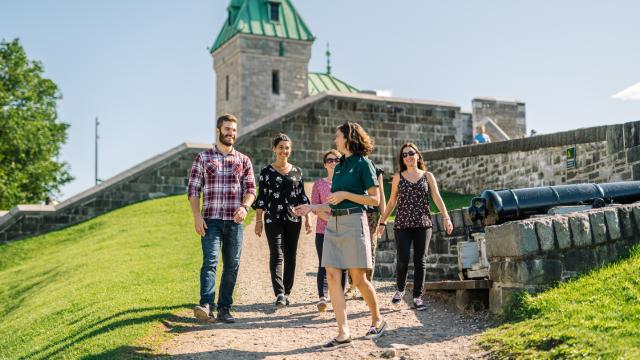 Visitors with a Parks Canada guide walk in front of a building at the Fortifications of Québec National Historic Site on a sunny day.