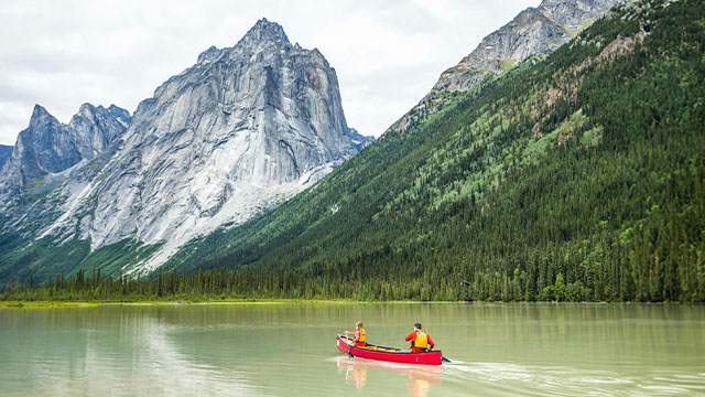 Two people canoeing on a green lake with rugged mountains in the background in Nahanni.