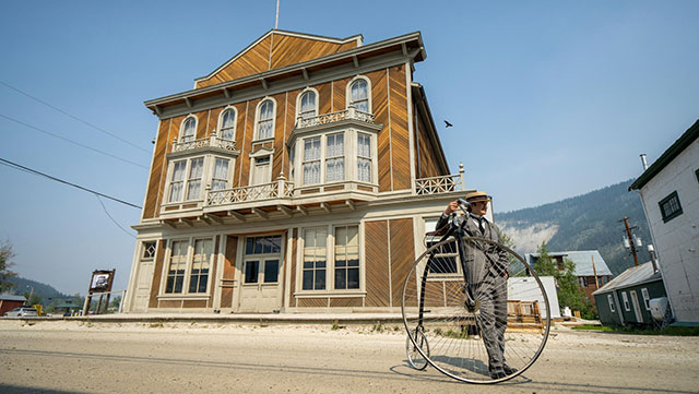 A man in period costume with a penny-farthing in front of heritage buildings in Dawson City.