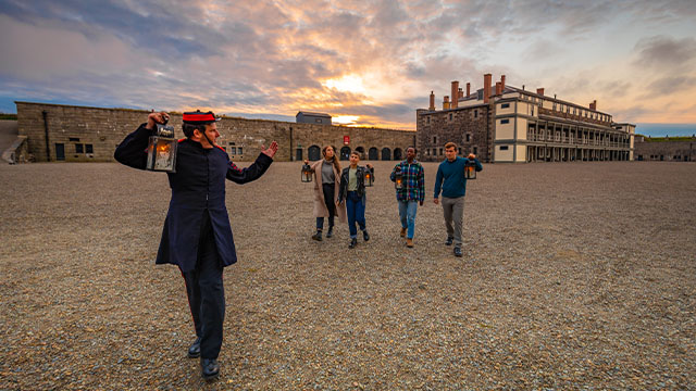 Four visitors and a guide with lanterns walk through the inner courtyard of the Halifax Citadel at dusk.