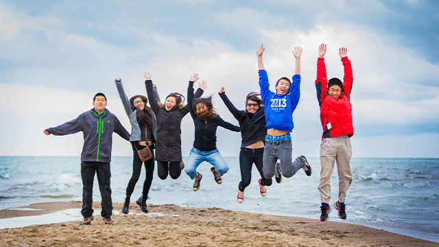 A group of young adults jumping at the Tip, next to in front of a lake, in Point Pelee National Park.