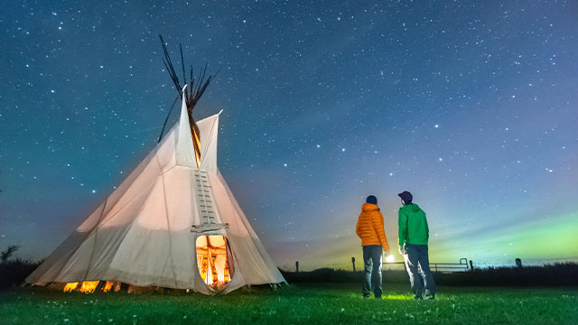 Two visitors gazing at the Big Dipper stars outside a tipi on a starry night at Rocky Mountain House National Historic Site.