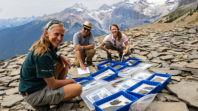 A Parks Canada guide shows fossil specimens with two hikers who are all sitting on rocks with mountains in the background of Yoho National Park.