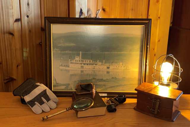 A framed, old, photo of a large boat, a workers glove, a magnifying glass, a lamp, a book, a lock, and other trinkets laid out on a table.