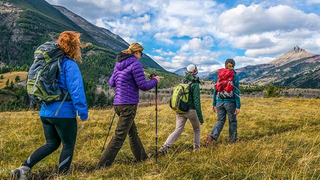 Four hikers traversing an open field with mountains in the background on a day with blue skies and clouds.