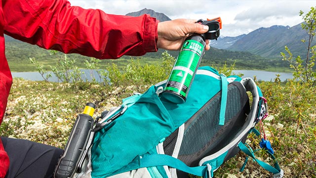 A person holds a bottle of spray over their backpack with a lake and mountains in the background.