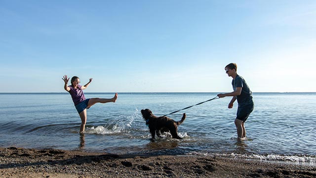 Two visitors play with their dog who is on a leash next to the water on a sunny summer day in Point Pelee.