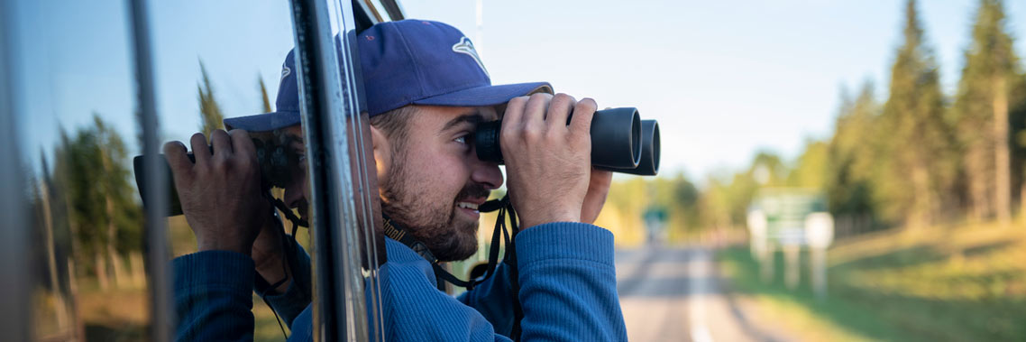 A man looks out the passenger seat window for wildlife with his binoculars along the Elk Island Parkway.