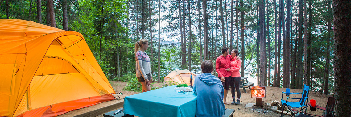 Four young adults are chatting around a picnic table with tents and a campfire in the background at La Mauricie National Park.