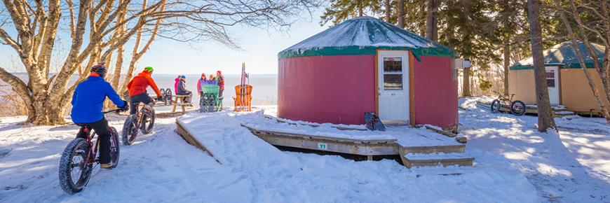 Un groupe d’amis avec des vélos, des raquettes et des skis près de deux yourtes sur le bord de la baie de Fundy par une journée d’hiver ensoleillée.