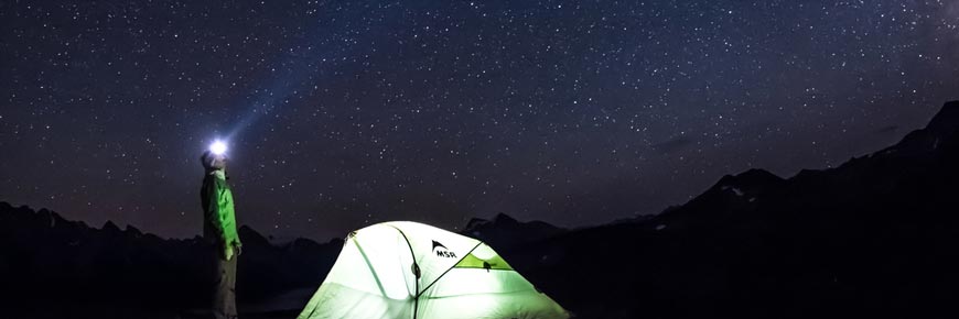 A man with a headlamp gazes up at the stars in front of his tent.