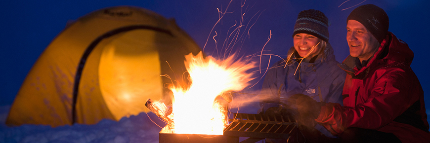 Deux adultes sont assis au bord du feu près de leur tente lors d’une soirée d’hiver.