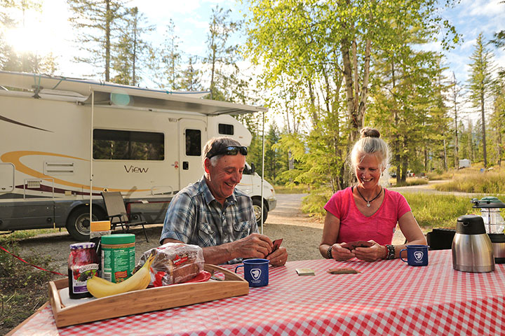 Two adults play cards on a picnic table in front of their RV with their campsite in the background