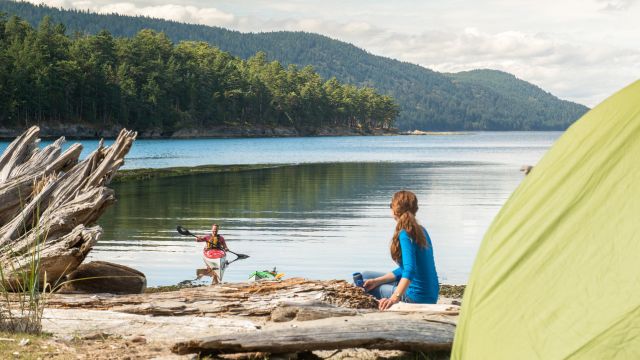 A kayak heads toward an adult and a tent on the shoreline, with coastal mountains in the background in Gulf Islands.