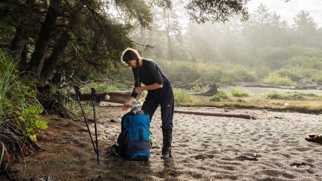 A hiker bends over a backpack beside hiking poles on a lush beach in a sunny haze in Pacific Rim.