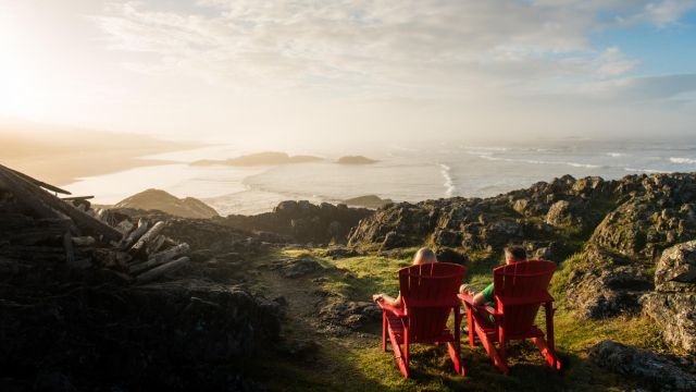 Two adults in red chairs look out at a magestic coastal view with grassy rocks and strong waters meltiung into the skyline.