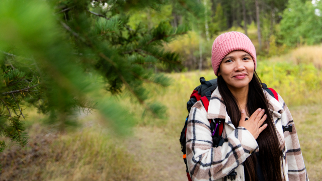 Woman standing in a forest clearing, facing forward among tall trees.