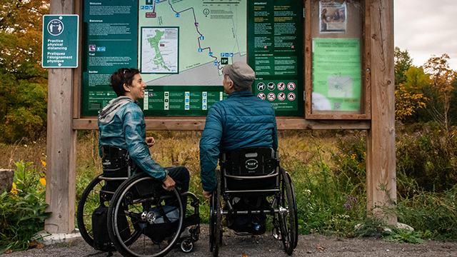 Deux personnes en fauteuil roulant devant un panneau à l’entrée d’un sentier au parc urbain national de la Rouge.