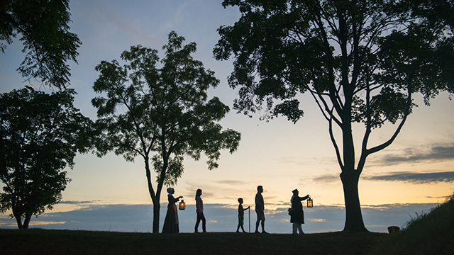 Shadows of 5 people in historic outfits carry lanterns and walk in a line in between trees