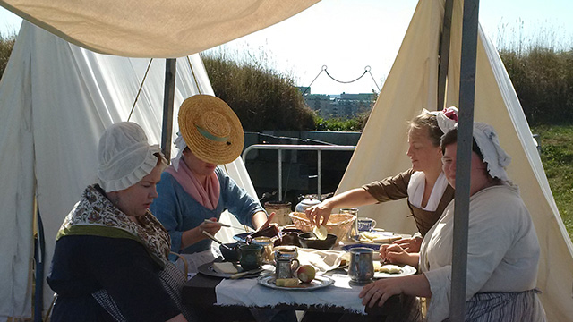 Four women in 18th-century costume take part in a re-enactment of a military camp, sitting down to eat