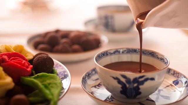 Hot chocolate being poured into a tea cup on a table with food in the background